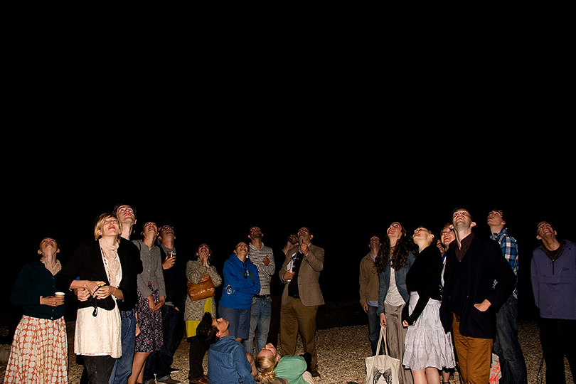 A large group of people standing in the dark on a pebble beach, illuminated only by the flash of the camera, are all looking upwards towards a spot in the dark black sky, as if watching something happen.