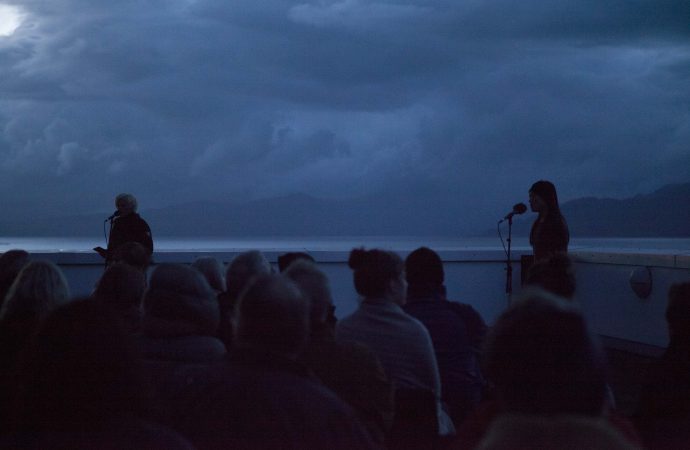At dawn a group of people watch two performers speaking into microphones. Behind them is a wide expanse of sea, with cloud covered mountains in the distance. The dawn light creates dark shadows and casts everything in a pale blue hue.