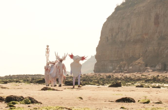 A line of people, dressed in bright white costumes resembling coral or strange sea creatures, walk across a sandy beach on a bright, hazy day. The beach is littered with stones covered in seaweed, and behind the procession is a large sheer cliff of stone.