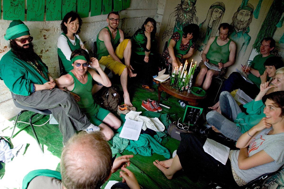 A group of people, all dressed in green items of clothing, sit in a circle in a small white room that is filled with green items such as; a large green rug, lit candles standing in green beer bottles, and green artworks drawn on and pinned to the walls.